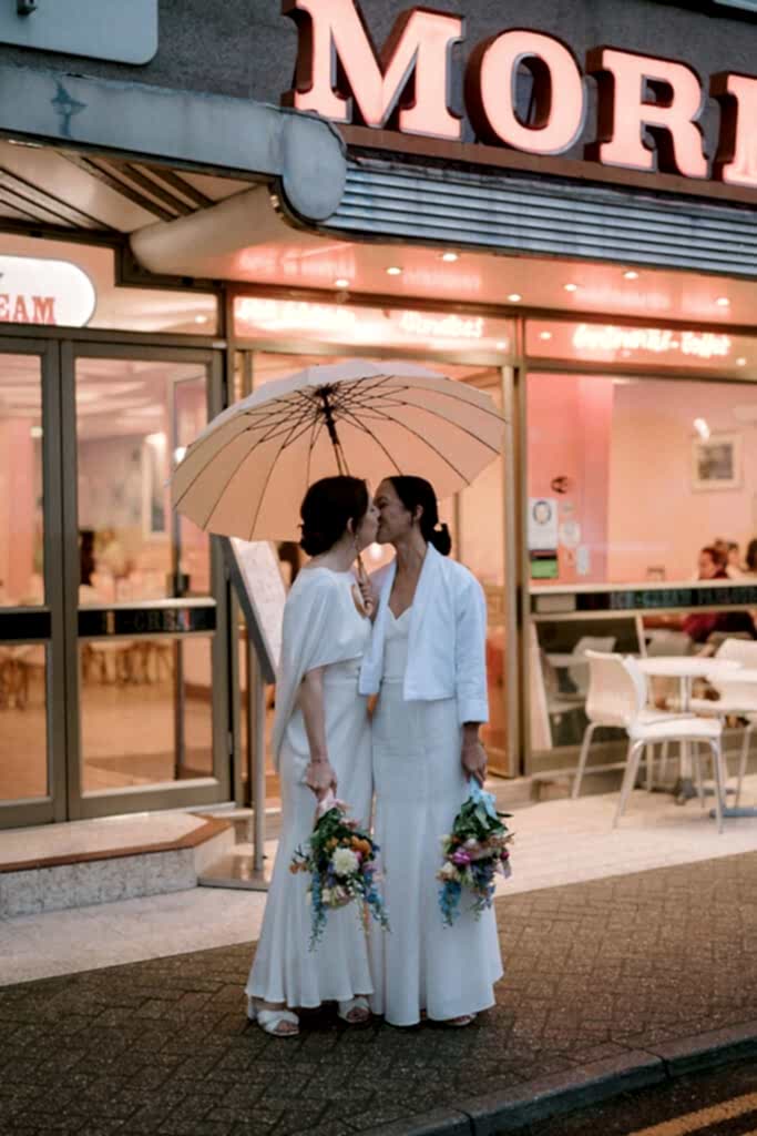 Two women in white wedding outfits share a kiss under a beige umbrella outside a retro-style ice cream shop in Broadstairs. Each holds vibrant flower bouquets, standing on the sidewalk with a neon sign glowing above them, capturing the essence of their love and celebration. Image by Pearce Wedding Photography.