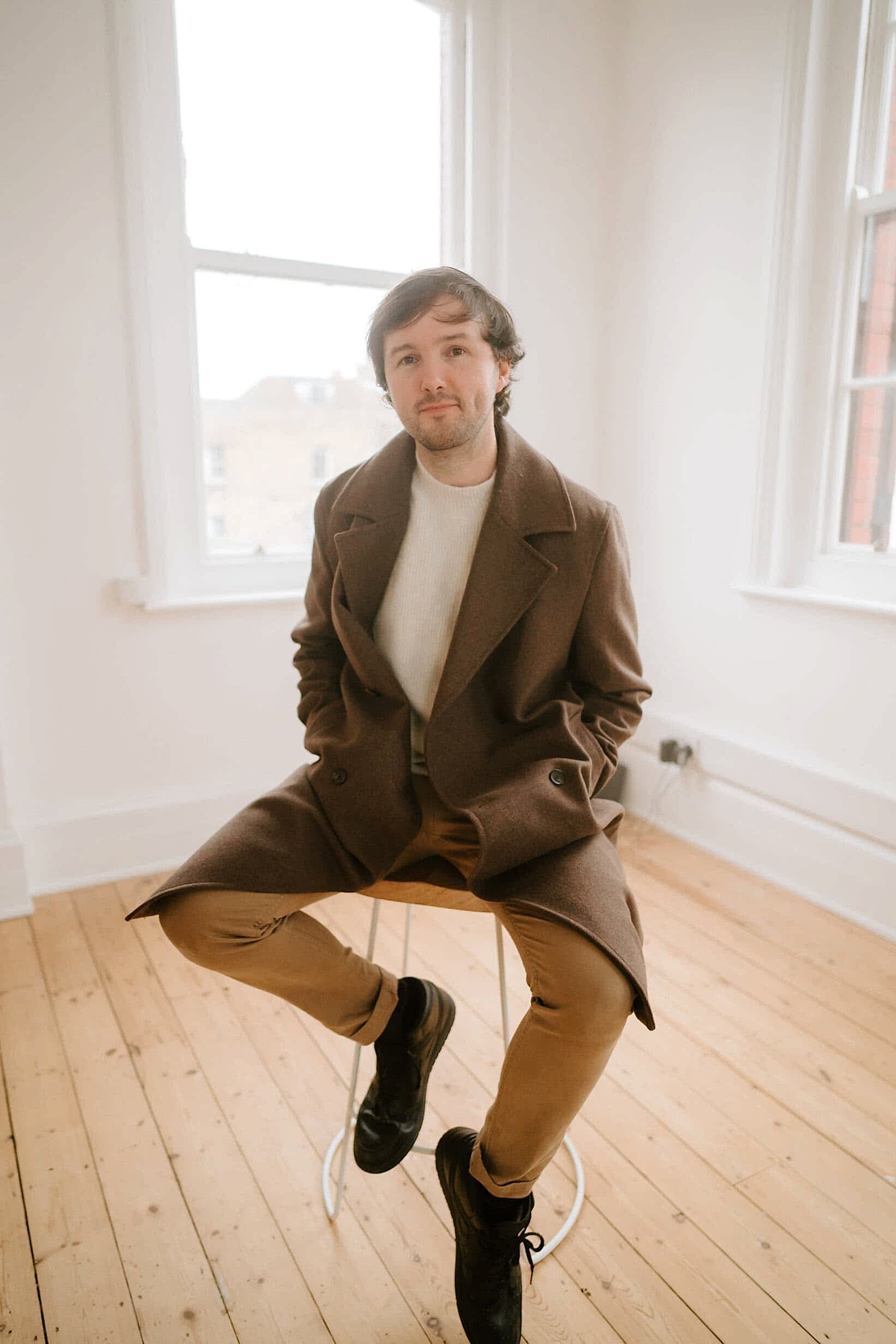 A person sits on a stool in a bright room with wooden flooring, reminiscent of a studio. Dressed in a brown coat, beige pants, and black shoes, they appear ready for their session with the Kent wedding photographer. Light streams through two large windows, casting soft shadows around them. Image by Pearce Wedding Photography.